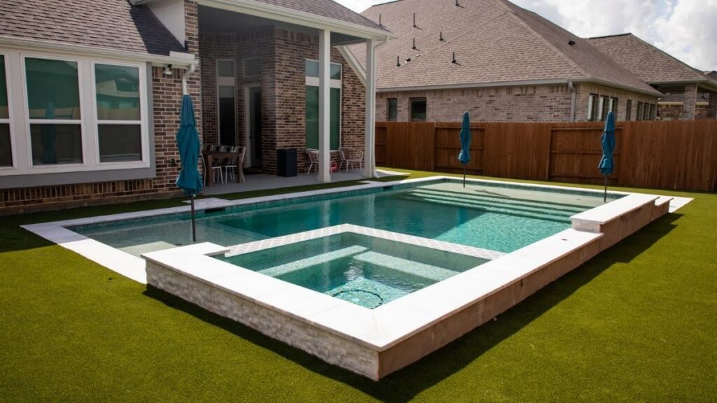 "Rectangular pool with raised spa and white stone coping surrounded by artificial turf behind a brick new build home with a covered patio"
