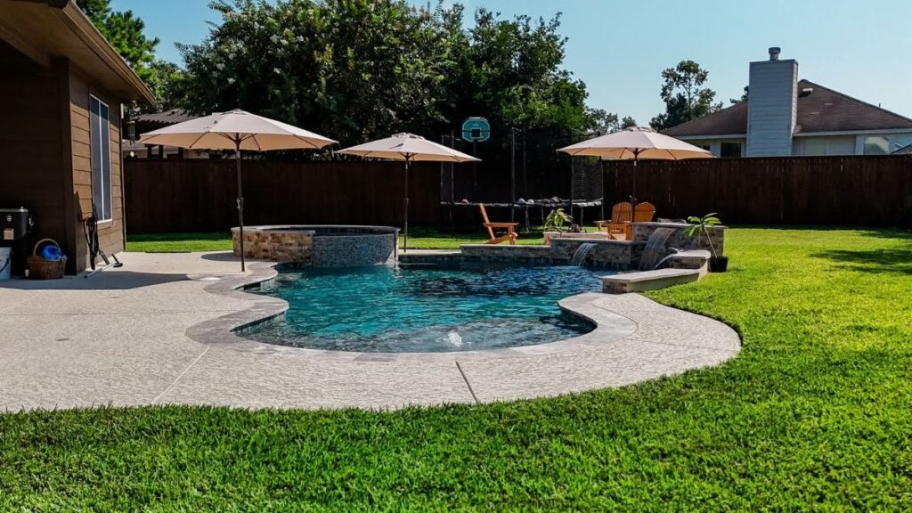 "Freeform pool with raised spa, stone waterfall features, and textured concrete deck in a fenced backyard with patio umbrellas and lawn"