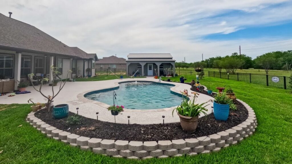 "Freeform inground pool with a textured concrete deck, landscaped planters, and a pool house in a North Texas backyard"