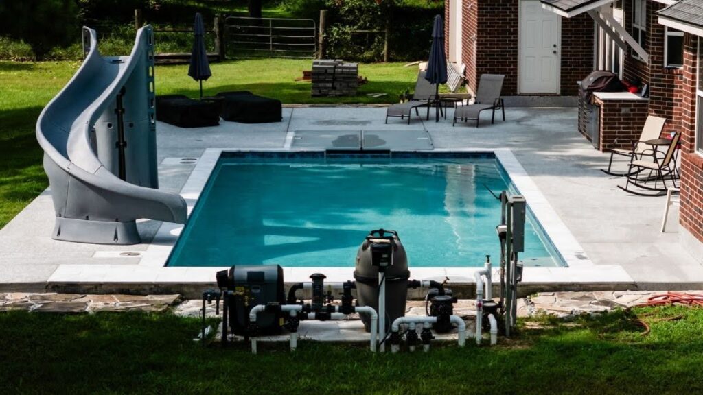 "Rectangular backyard swimming pool with a gray spiral slide, pool pump and filter equipment, lounge chairs, and an outdoor kitchen beside a brick house."